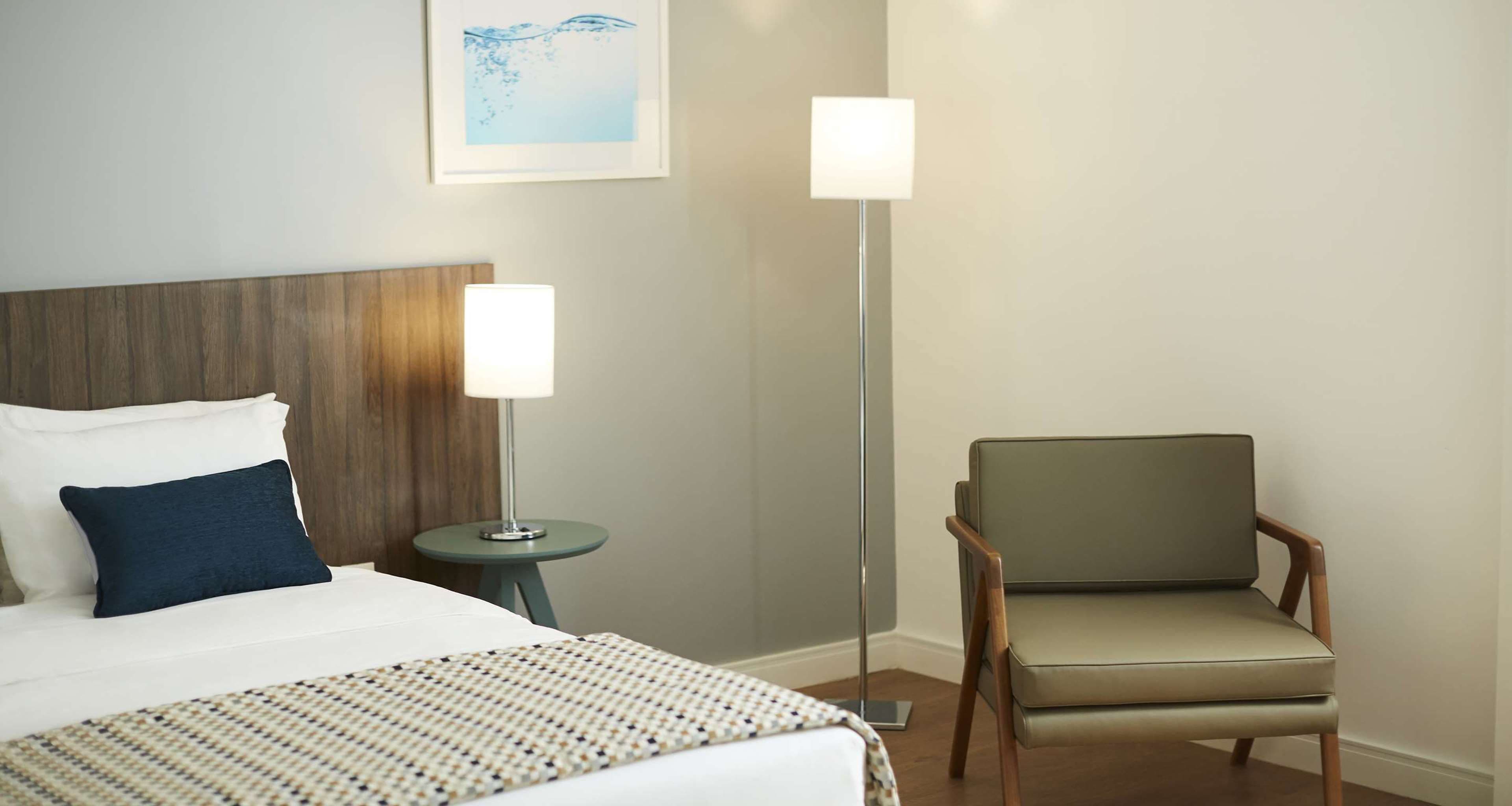 A hotel room with a bed, a chair, a bedside table, and a floor lamp.