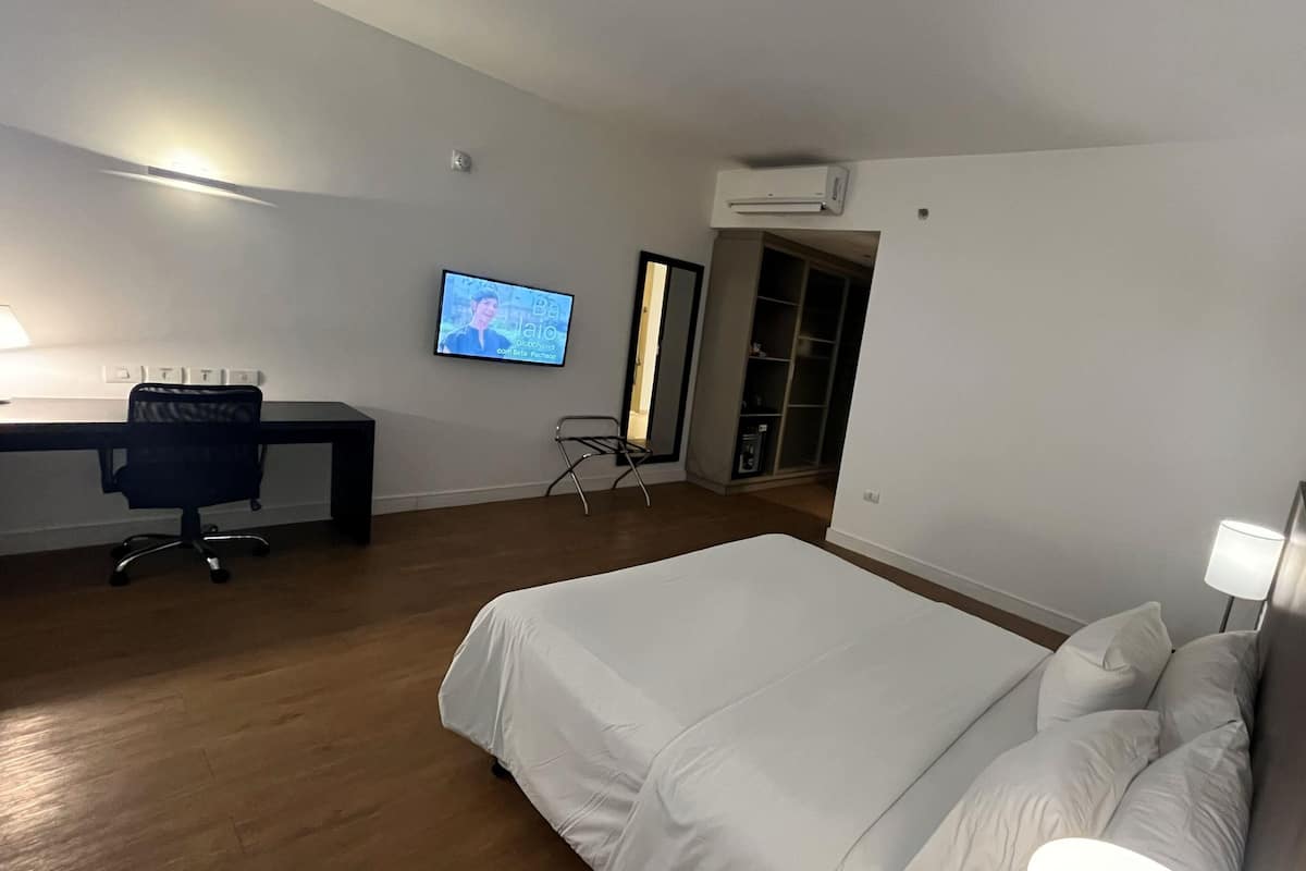A modern hotel room with a large bed, a desk with a computer, a TV, and an air conditioning unit.