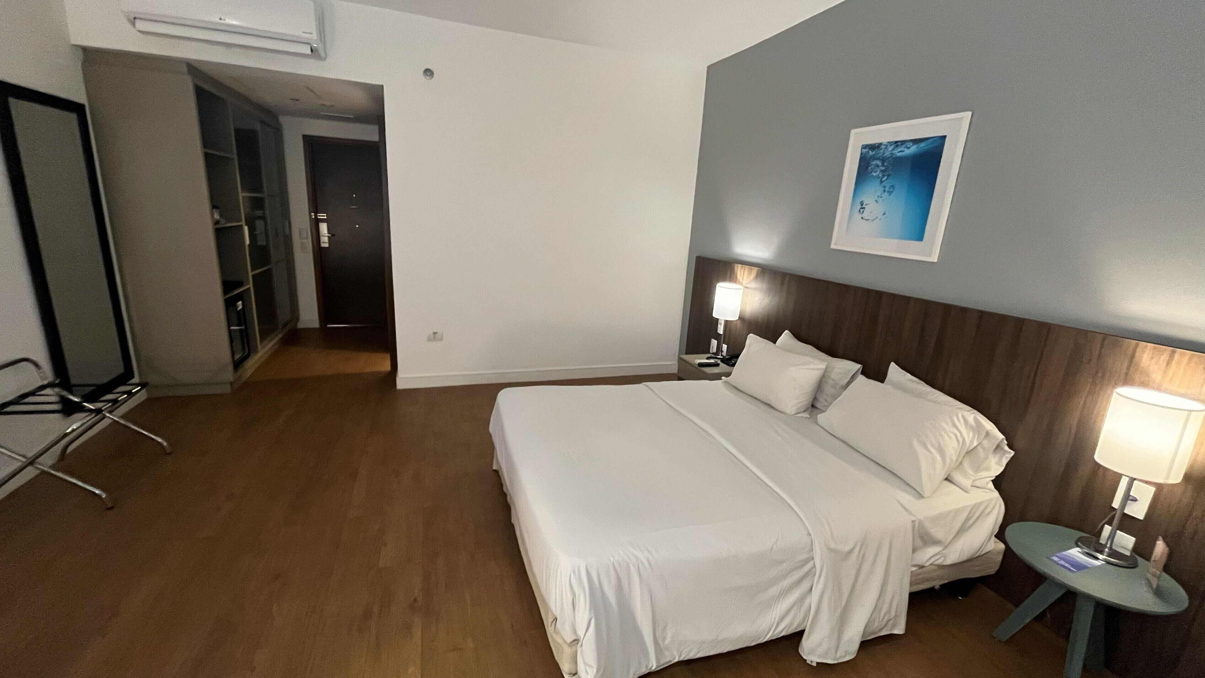 Une chambre d’hôtel avec un grand lit, des tables de chevet avec des lampes, une tête de lit en bois, un tableau encadré au mur et une porte donnant sur un couloir.