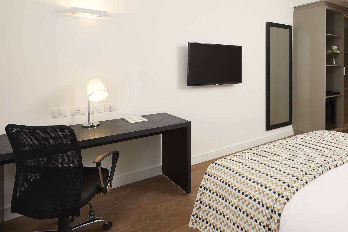 A hotel room with a bed, a desk with a chair, a TV, a mirror, and a wardrobe.