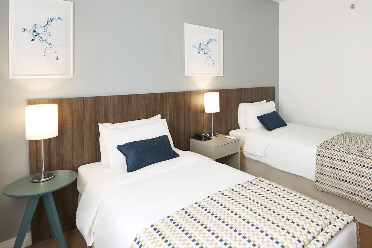A hotel room with two beds, a nightstand with a lamp, and artwork on the wall.