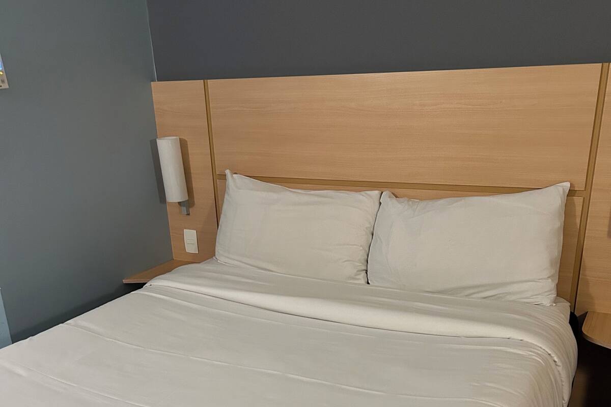 A neatly made bed with white linens, a wooden headboard, and a wall-mounted light fixture.