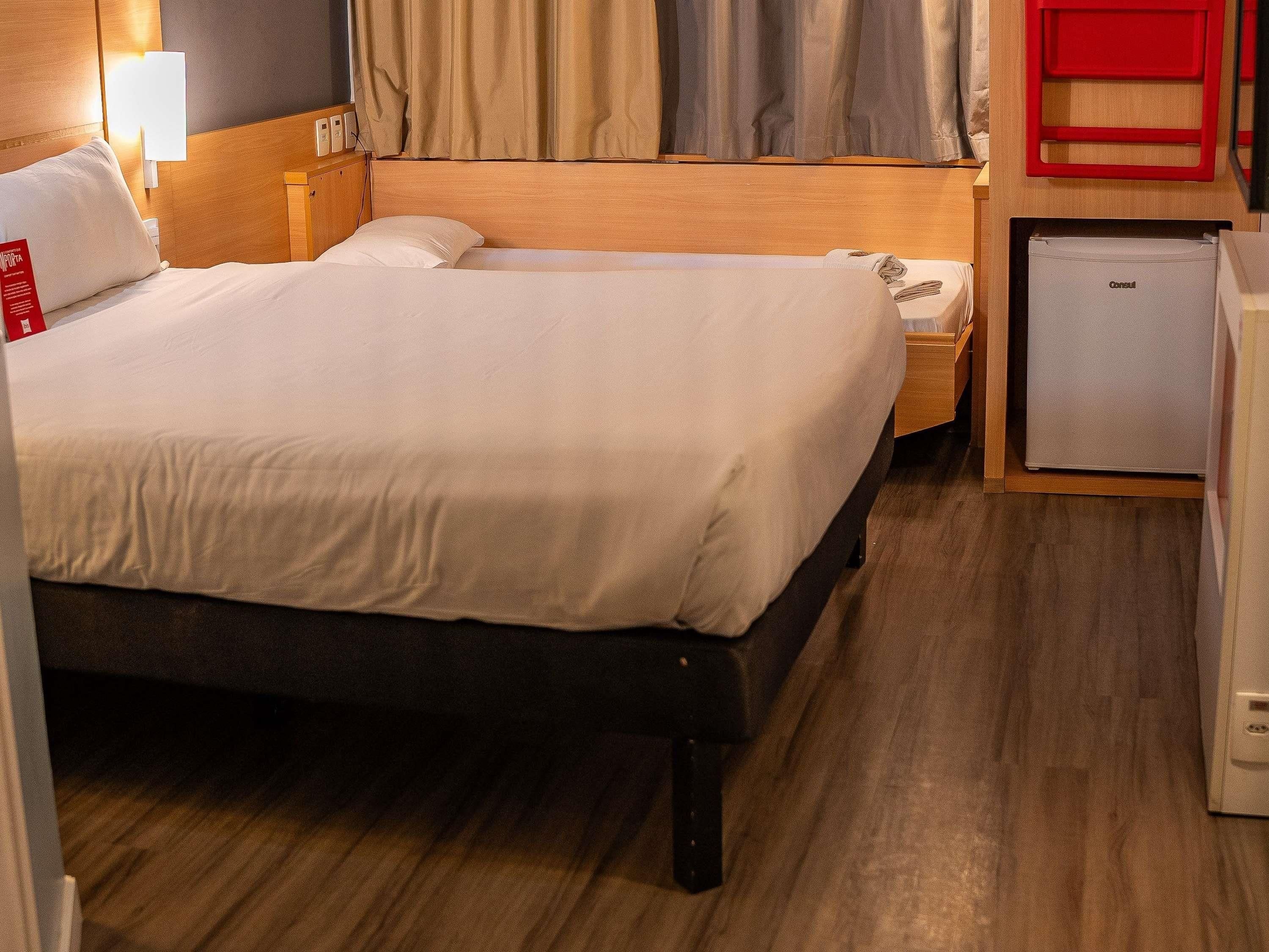 Un lit double avec une tête de lit en bois, une table de chevet avec une lampe et un petit réfrigérateur.