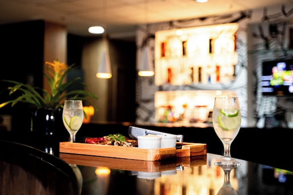 Zona de bar con una mesa de madera, dos vasos de una bebida a base de lima, un pequeño plato con comida y una planta en maceta.