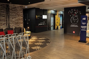 Um saguão de hotel com balcão de recepção, área de estar e parede com diversos avisos e obras de arte.