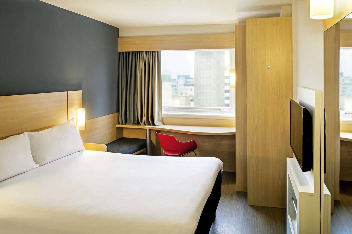 A hotel room with a large bed, a desk, a TV, and a window with a city view.