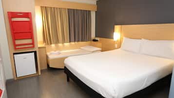 A hotel room with a large bed, a bedside table, a small fridge, a red object on the wall, and a window with curtains.