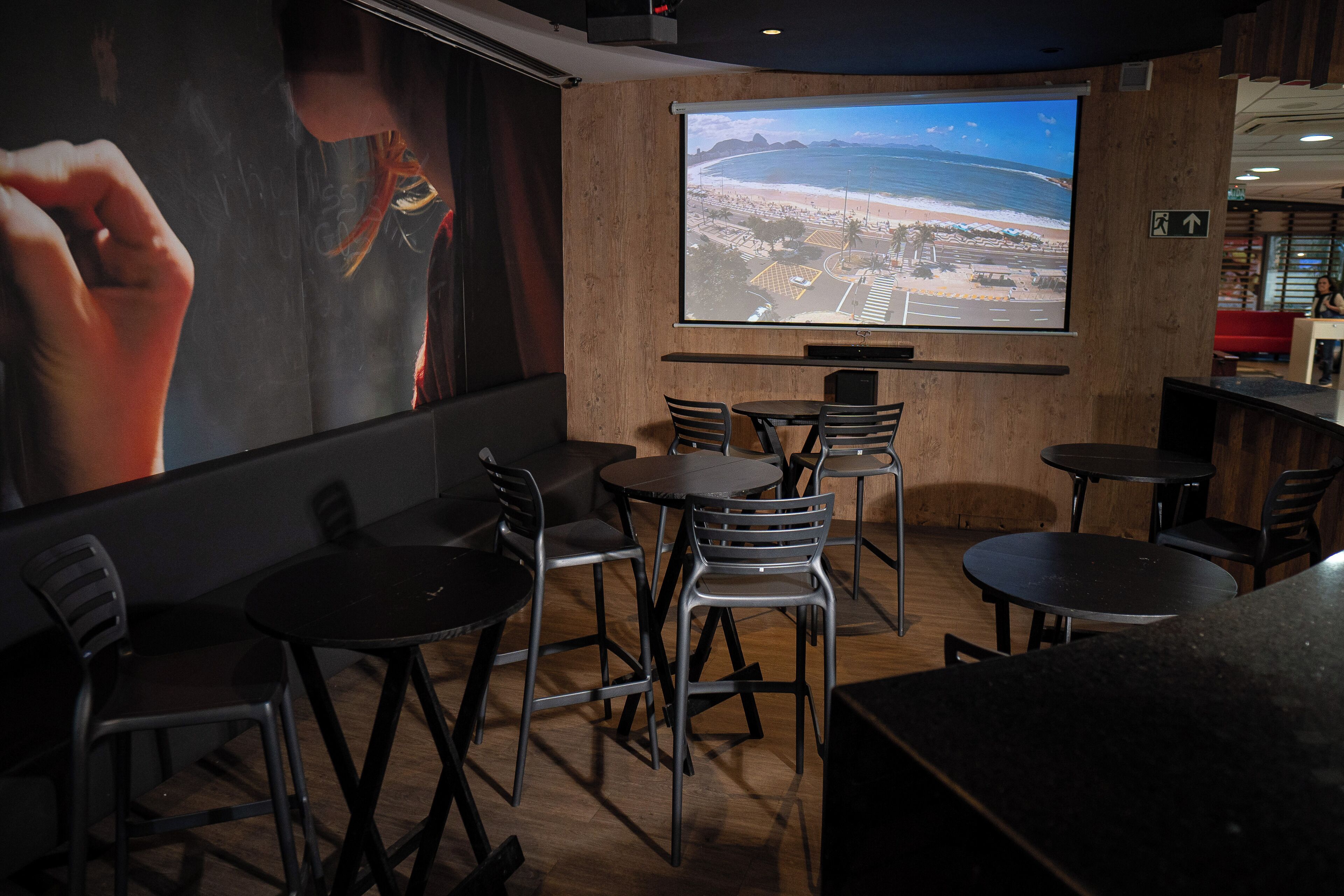 Ein Restaurant mit großem Bildschirm, der einen Strand zeigt, mehreren Tischen und Stühlen sowie einer Holzvertäfelung.