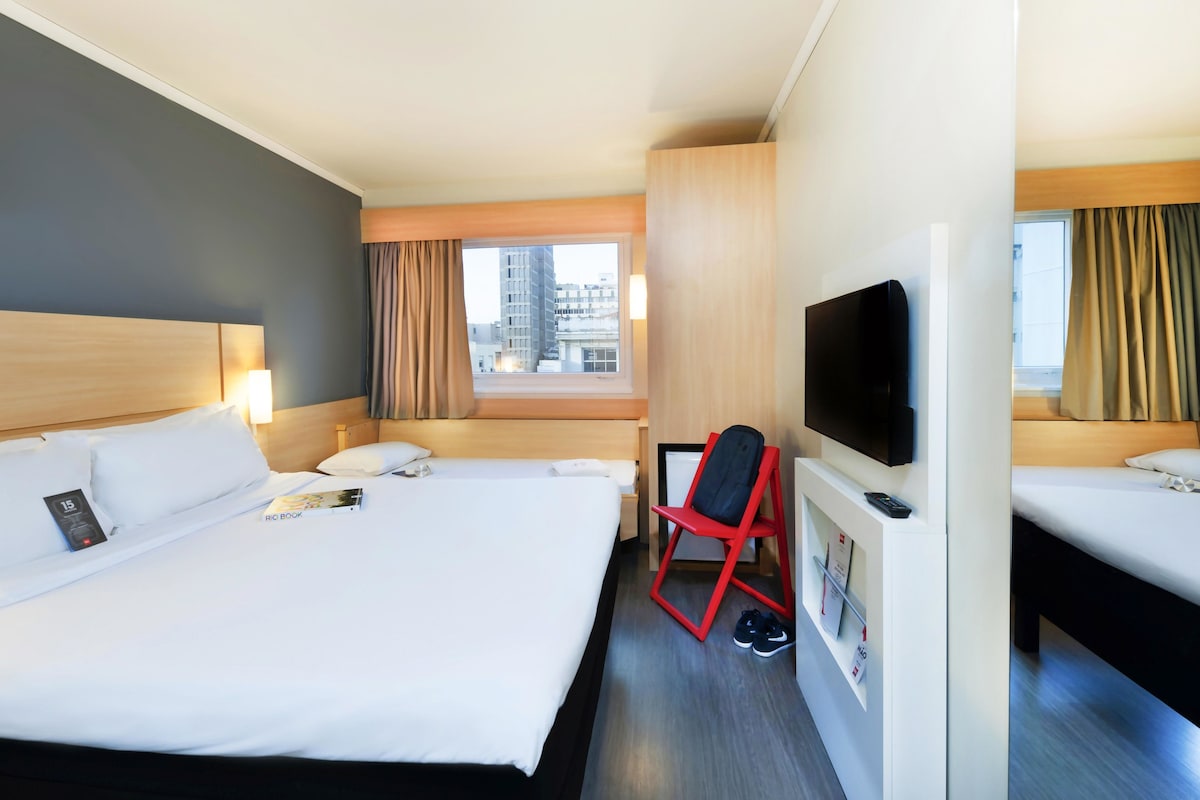 A hotel room with a large bed, a TV, a desk with a chair, and a window with curtains.