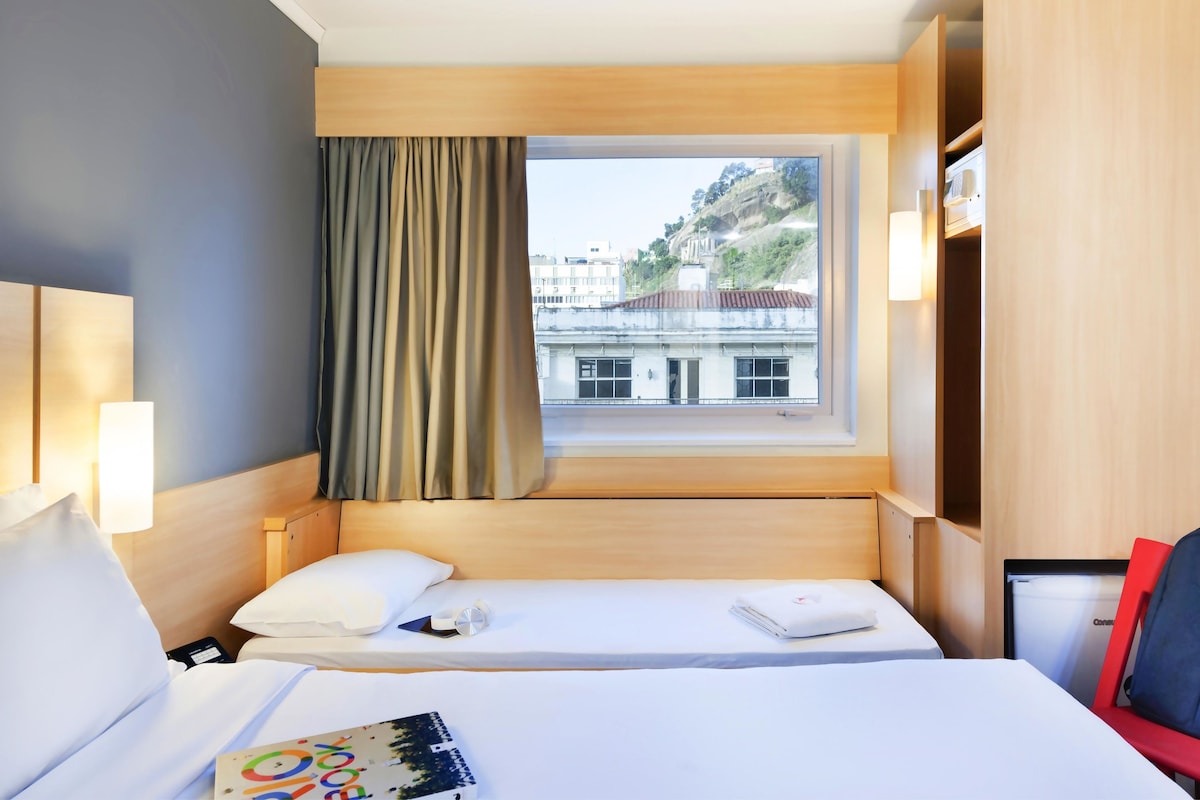 Habitación de hotel con cama, una silla, escritorio, lámpara, ventana con cortinas y vistas a edificios.