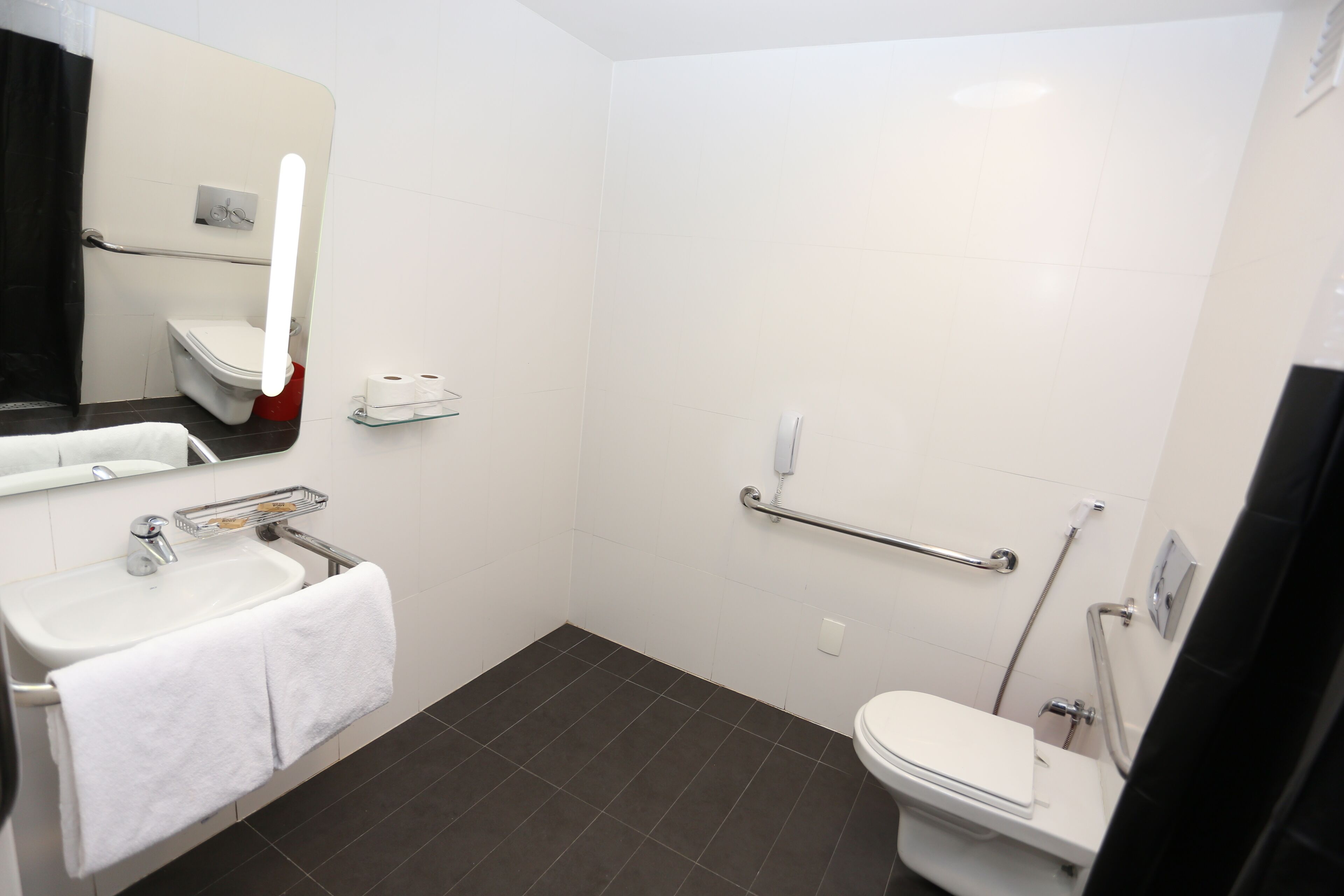 Un baño moderno con lavamanos, espejo y retrete blancos. Se observa una barandilla y un estante de vidrio con artículos de tocador.