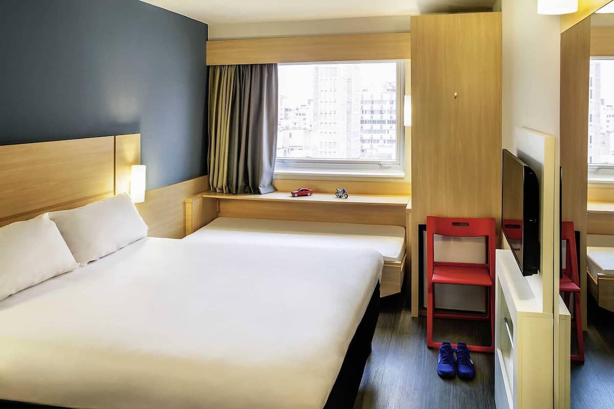 A hotel room with a large bed, a TV, a small desk, and a window with a view of buildings.
