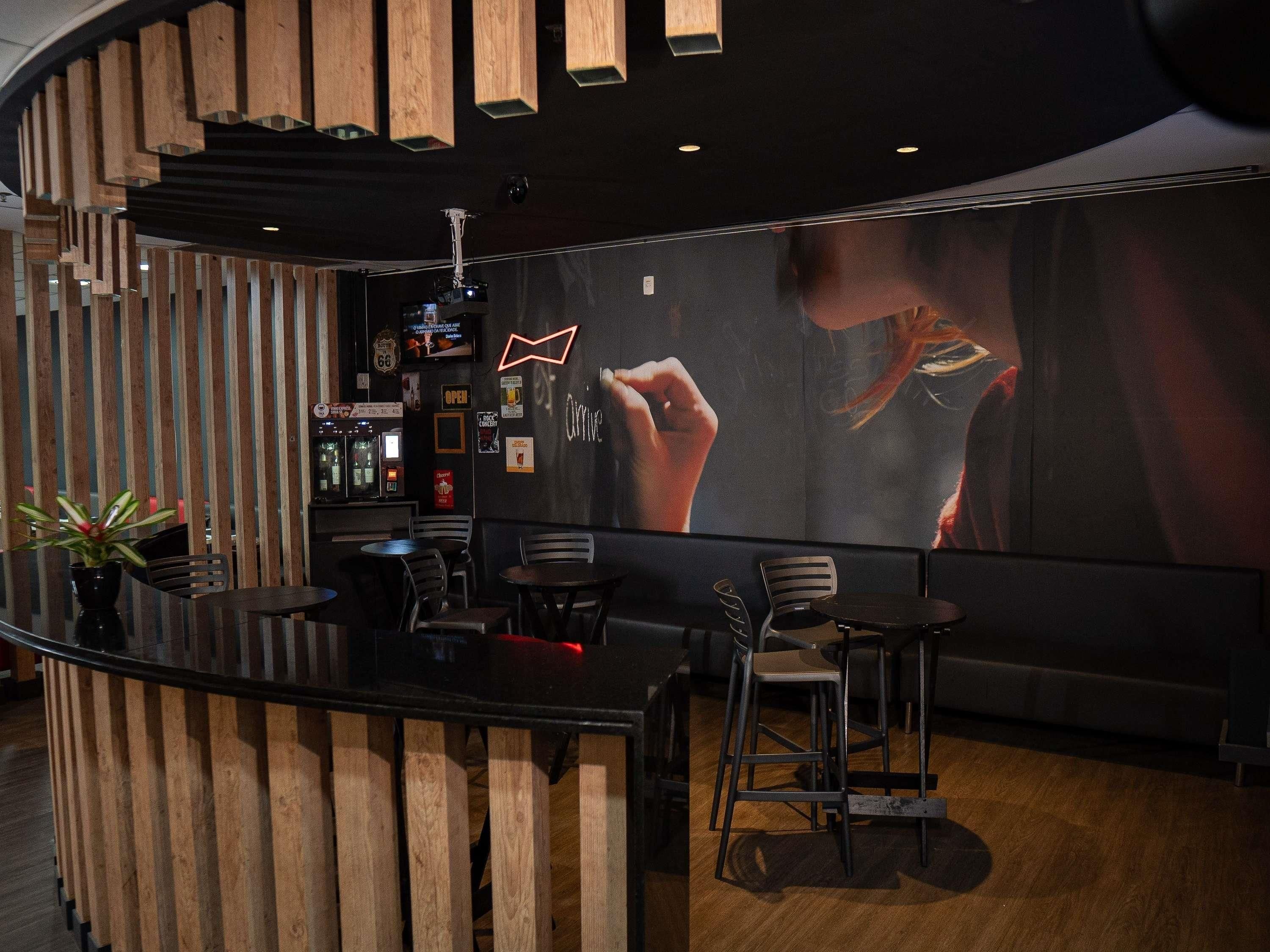 Zona de bar amb un taulell de fusta, seients i una pantalla gran on es veu una persona.