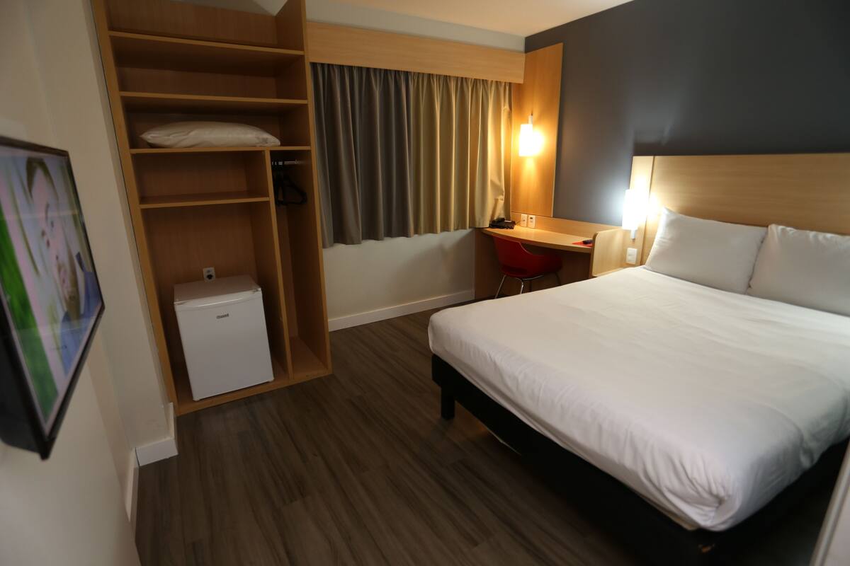 A hotel room with a bed, a desk, a wardrobe, and a small refrigerator.