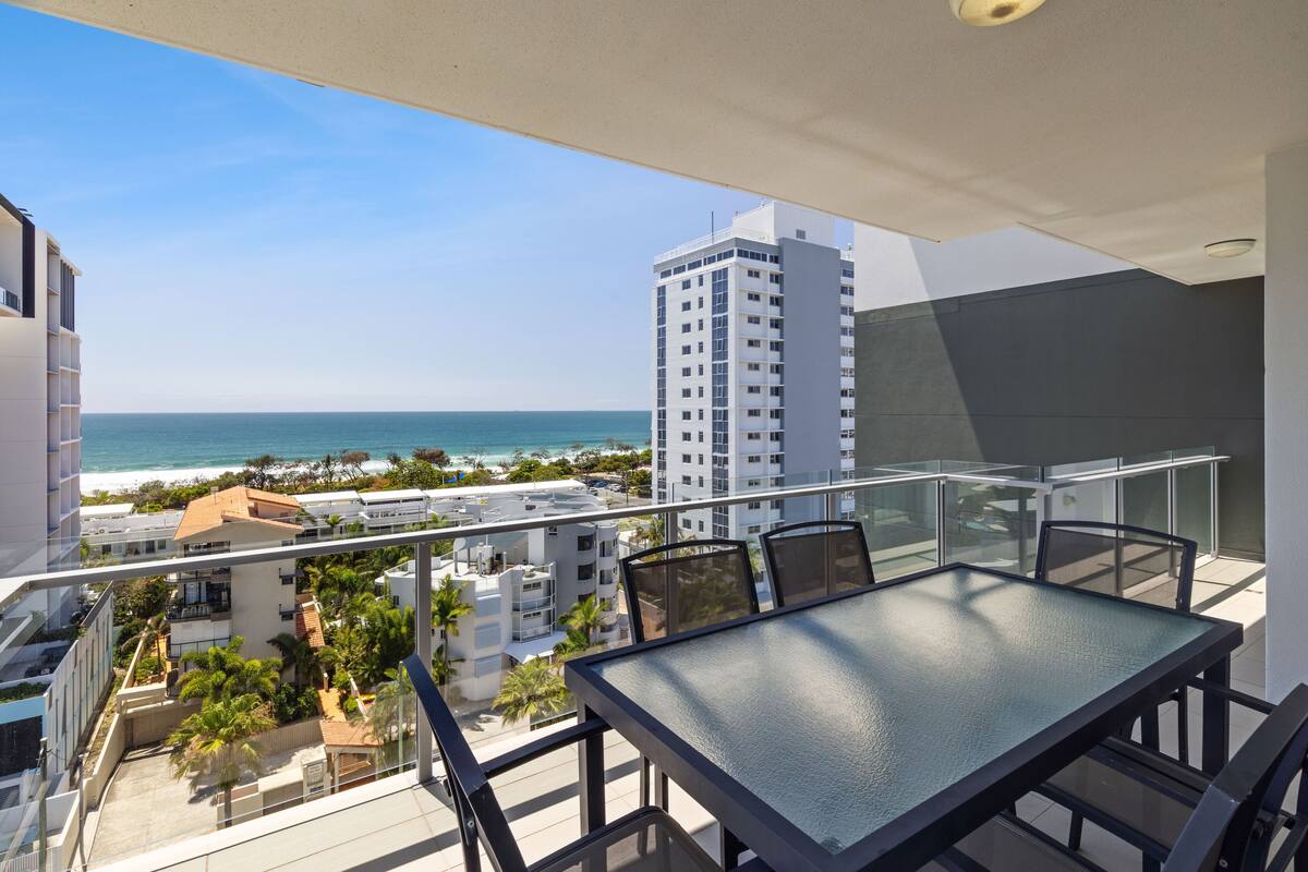 Un balcón con una mesa y sillas de vidrio, con vista a una playa y el océano.