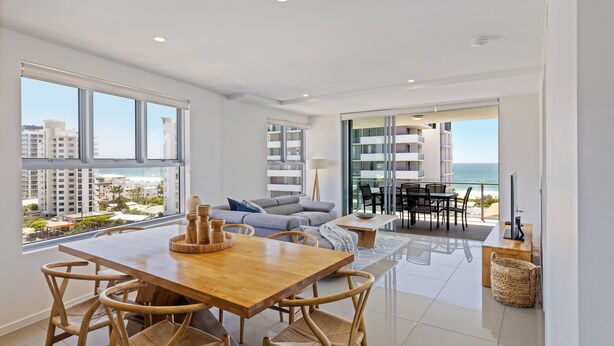 A modern living room with a wooden dining table and chairs, a sofa, and a view of the ocean. - Verve on Cotton Tree (Maroochydore)