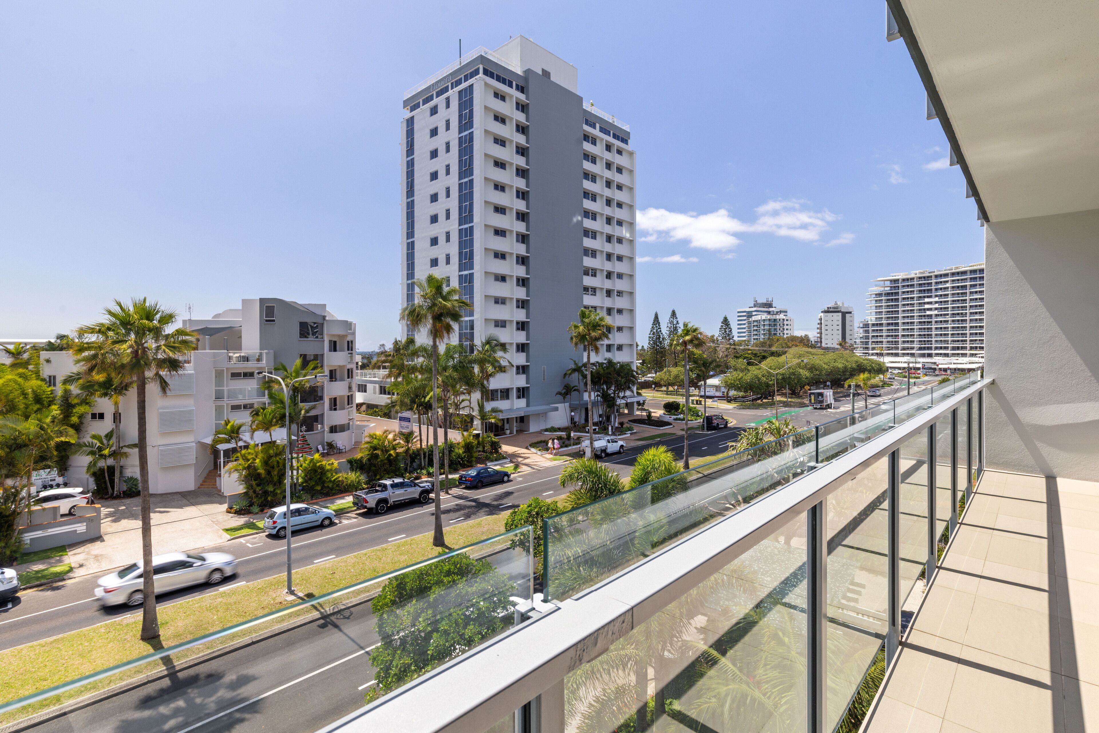 Un immeuble de grande hauteur au design moderne, doté d’un balcon en verre et offrant une vue sur une rue animée, avec des voitures et des palmiers.