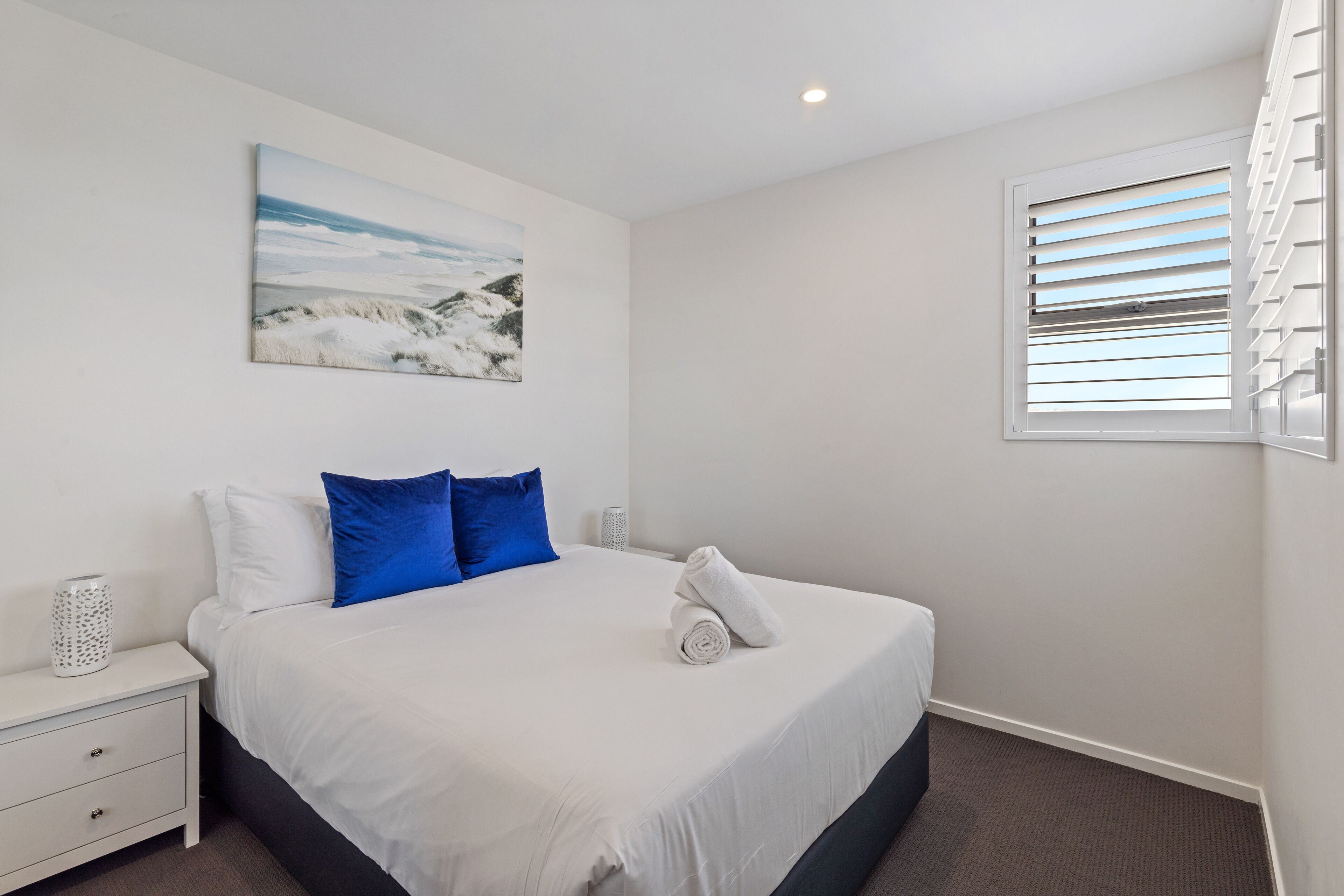 A neatly made bed with white and blue pillows, a rolled white towel, and a painting of a snowy landscape on the wall.