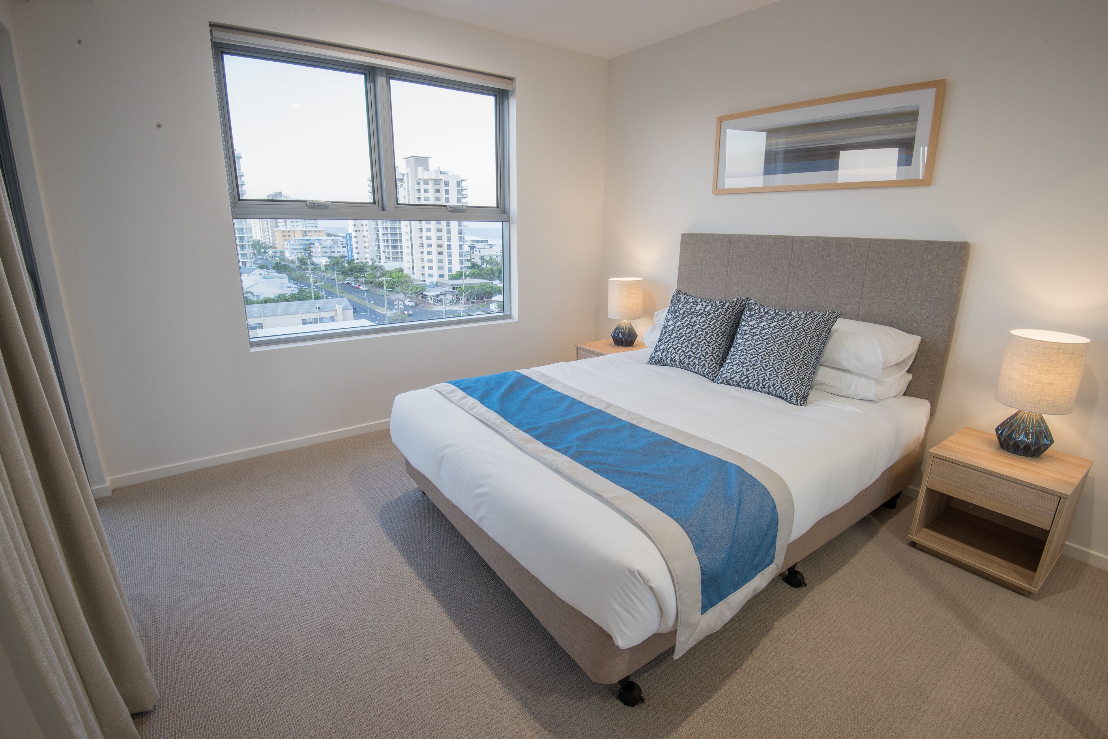 A hotel room with a large bed, two bedside tables, a window with a city view, and a framed picture on the wall.