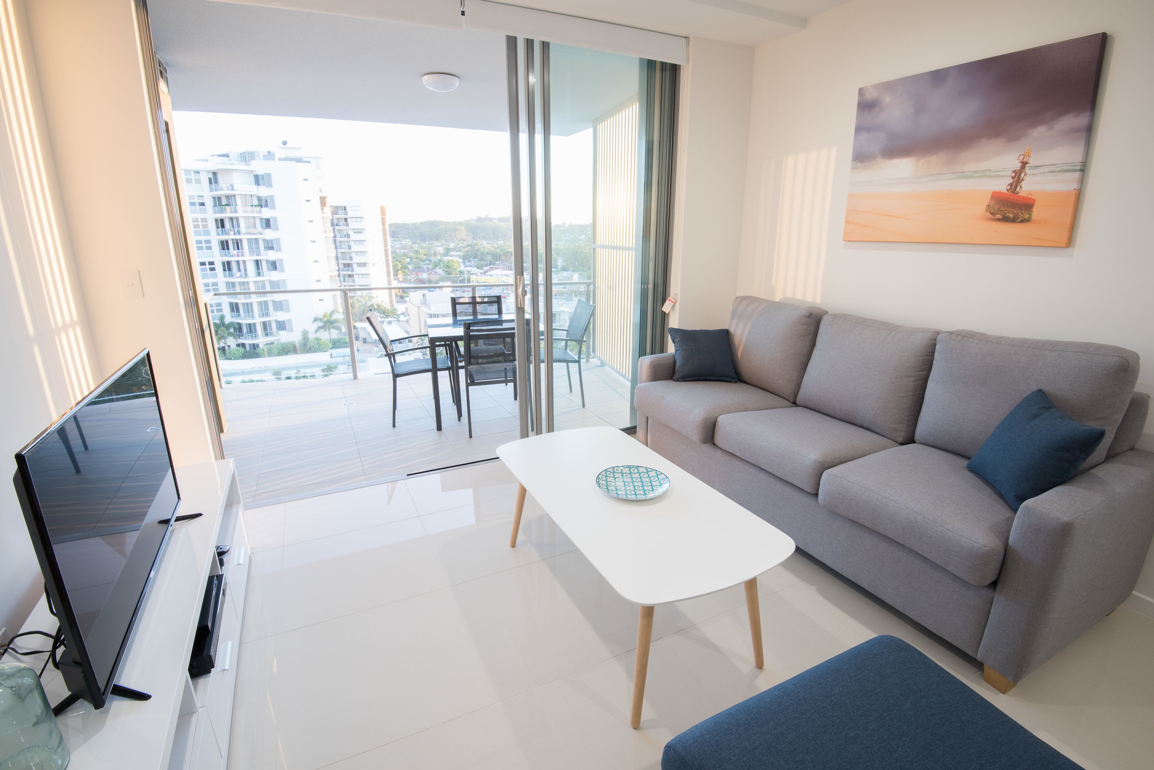 A modern living room with a flat-screen TV, a sofa, a coffee table, and a balcony with a view of buildings.