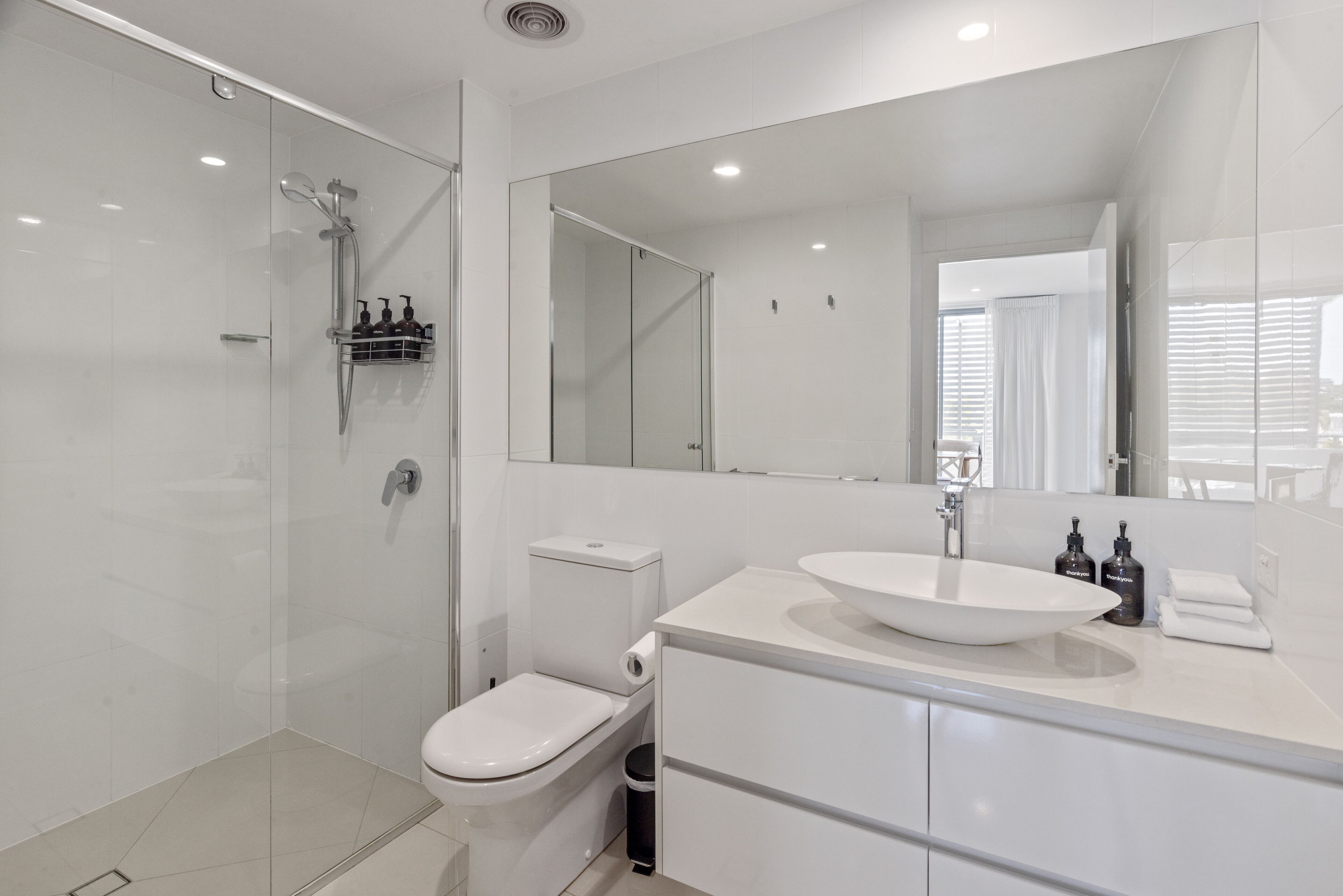 A modern bathroom with a glass-enclosed shower, a toilet, a sink with a large mirror, and a trash bin.