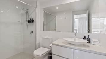 A modern bathroom with a glass-enclosed shower, a toilet, a sink with a large mirror, and a trash bin.