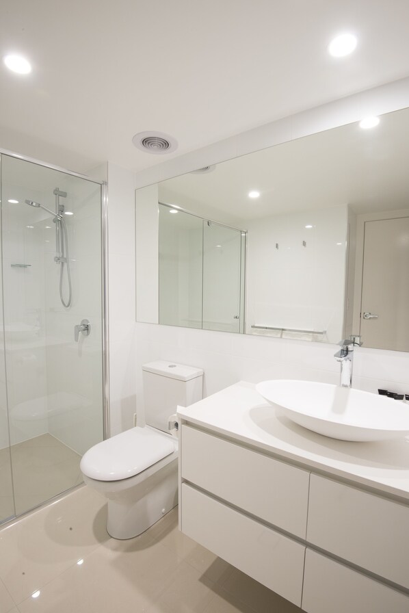 A modern bathroom with a glass shower enclosure, a white toilet, a large mirror, and a white vanity with drawers.