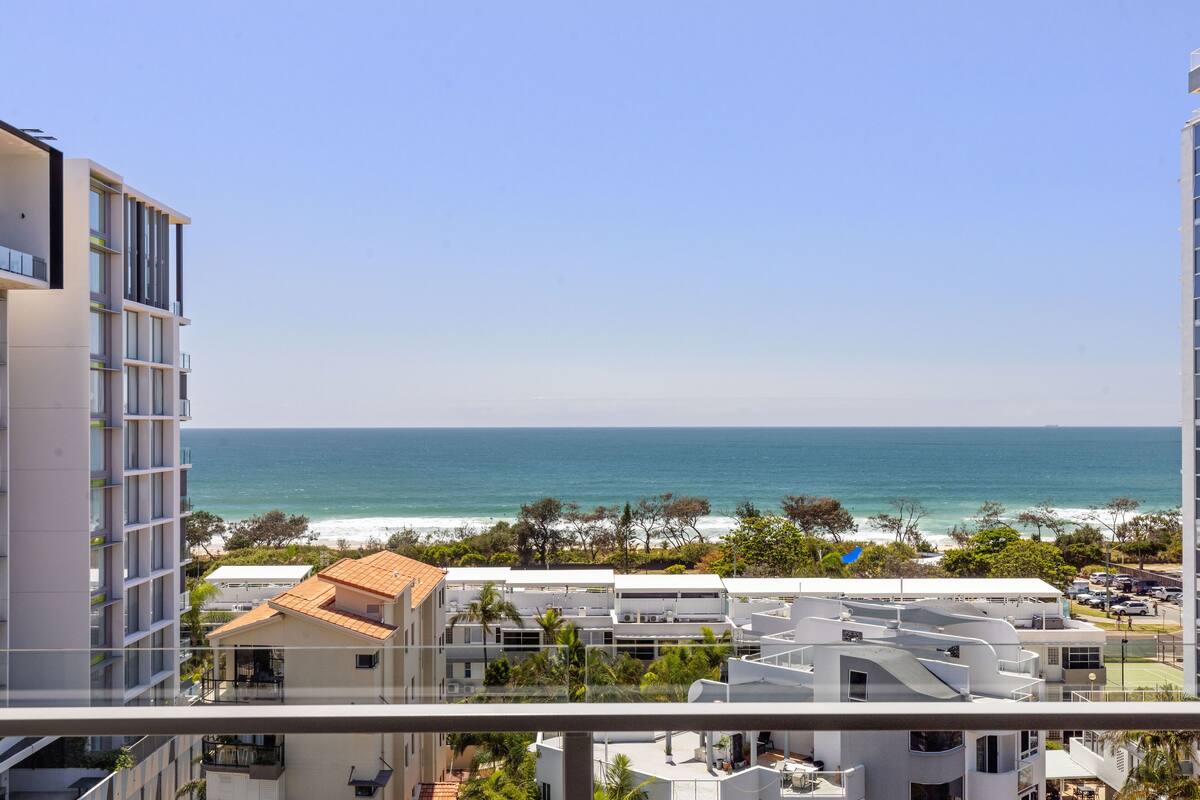 Vista costera desde un balcón con vistas a una playa, con edificios modernos y cielo despejado.