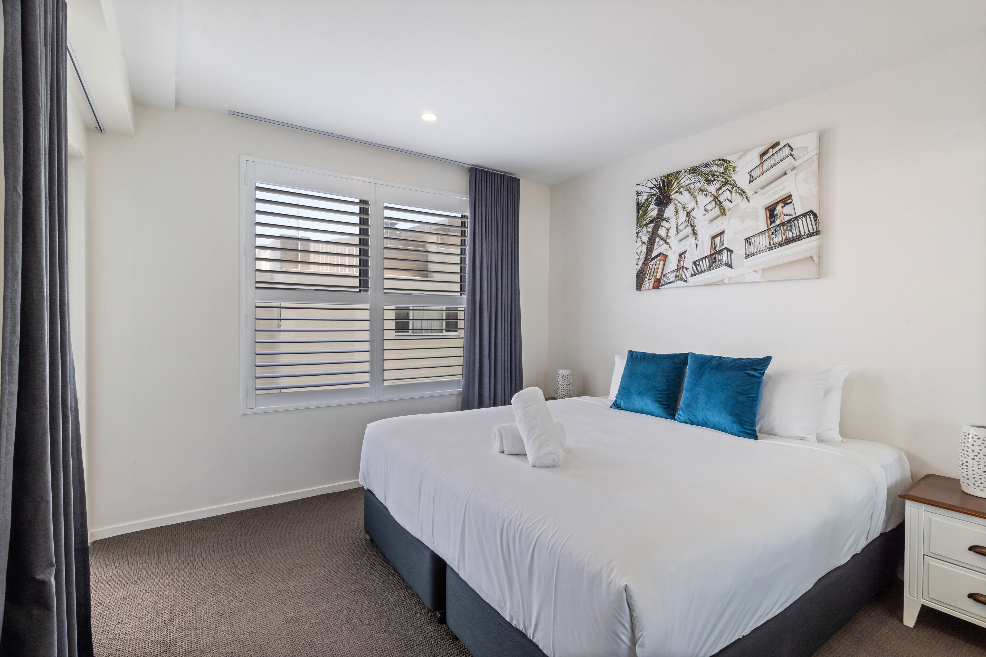 A hotel room with a large bed, a window with blinds, a nightstand, and a wall-mounted artwork.