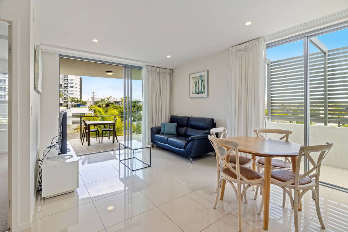 Una sala de estar moderna con sofá, mesa de comedor y sillas, televisor y un balcón con vistas a edificios y zonas verdes.