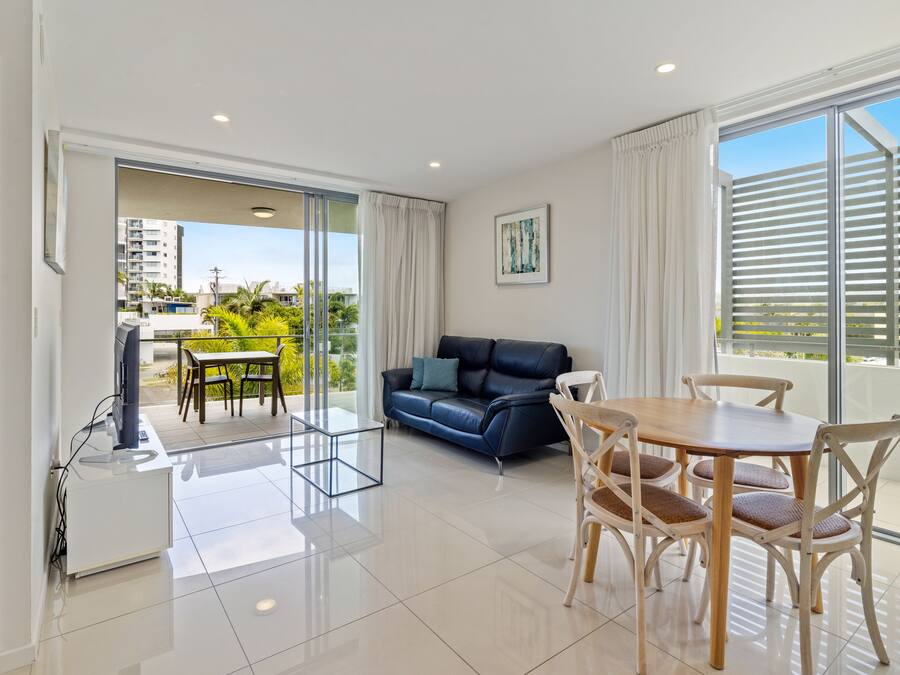 Un salon moderne comprenant un canapé, une table à manger et des chaises, un téléviseur, ainsi qu’un balcon offrant une vue sur les bâtiments et la verdure.