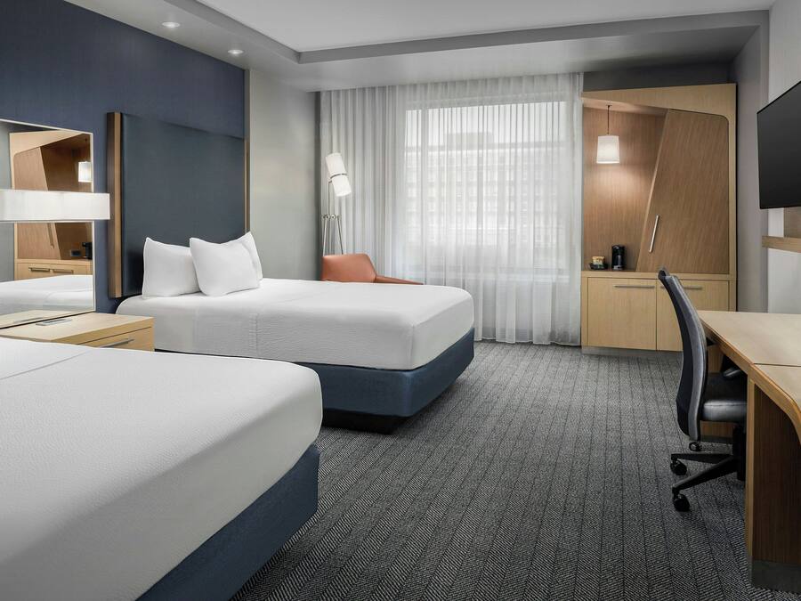A hotel room with two beds, a desk, a chair, a TV, and a large window with curtains.