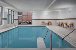 Una piscina con solárium empedrado, sillas y vista a un edificio de ladrillo.