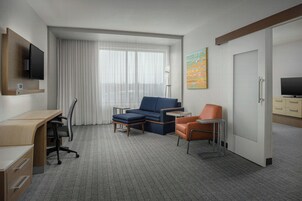 Habitación de hotel con escritorio, sofá azul, sillón naranja, televisor y un cuadro en la pared.