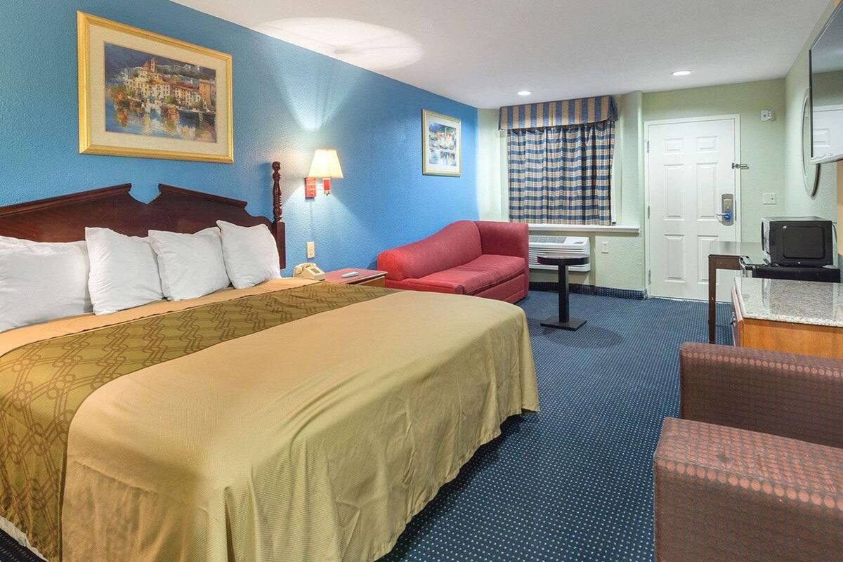 Quarto de hotel com cama, sofá vermelho, mesinha, luminária, televisão e janela com cortinas.