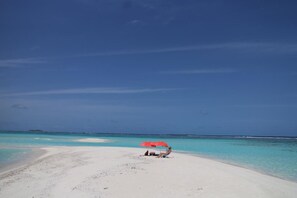 A beach with a person under a red umbrella, clear turquoise water, and a distant island.