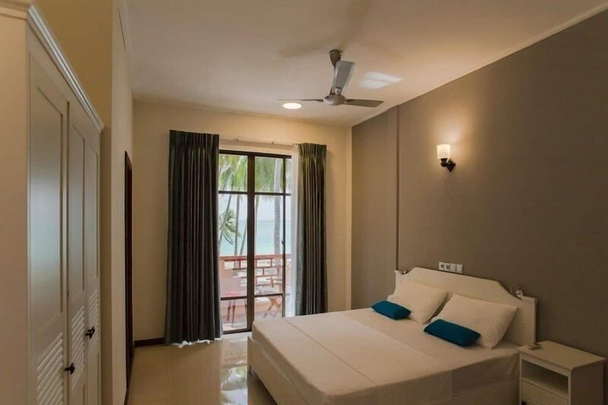A hotel room with a bed, a nightstand, a ceiling fan, and a view of the ocean through a large window.