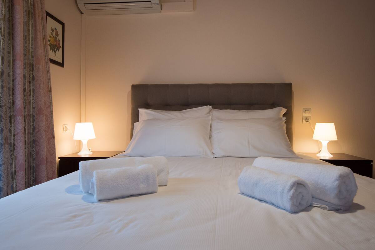 A neatly made bed with white linens and two rolled towels, flanked by two bedside lamps on a dark nightstand.