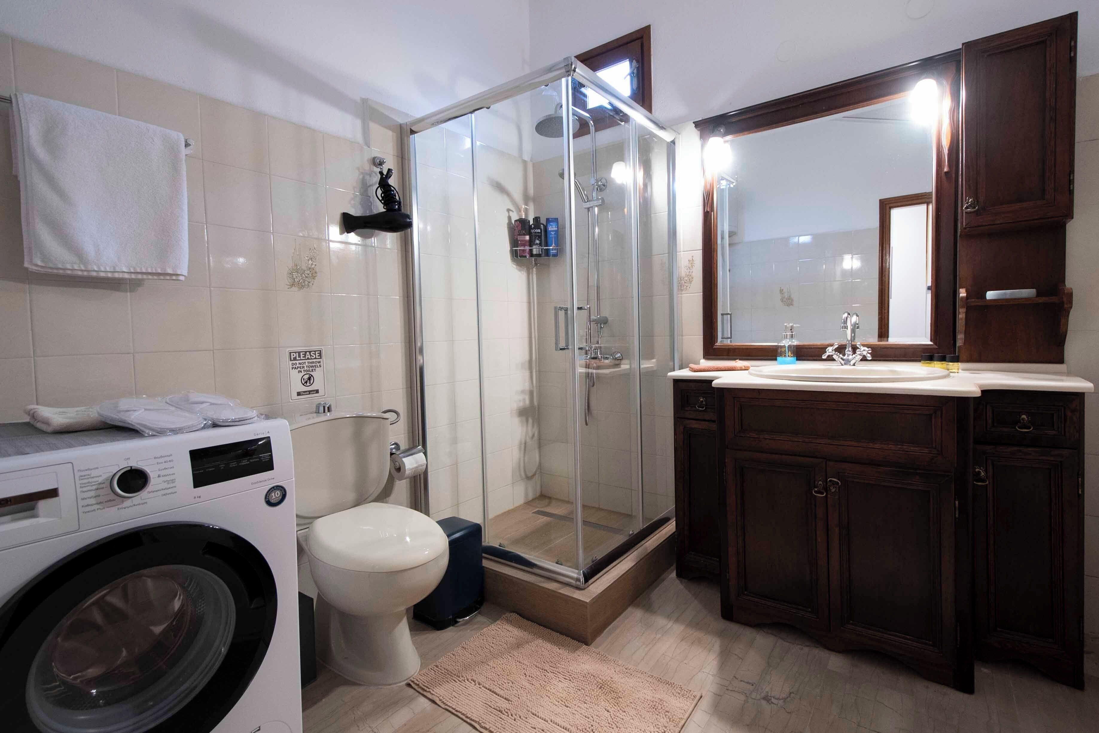 A bathroom with a glass-enclosed shower, a washing machine, a toilet, and wooden cabinets.