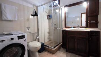 A bathroom with a glass-enclosed shower, a washing machine, a toilet, and wooden cabinets.