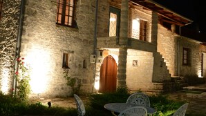 A stone building with a wooden door and windows, a paved area with a table and chairs, and a garden with plants. - Rodami (North Tzoumerka)