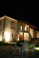 Un edificio de piedra con puerta y ventanas de madera, un área empavedada con mesa y sillas, y un jardín con plantas.