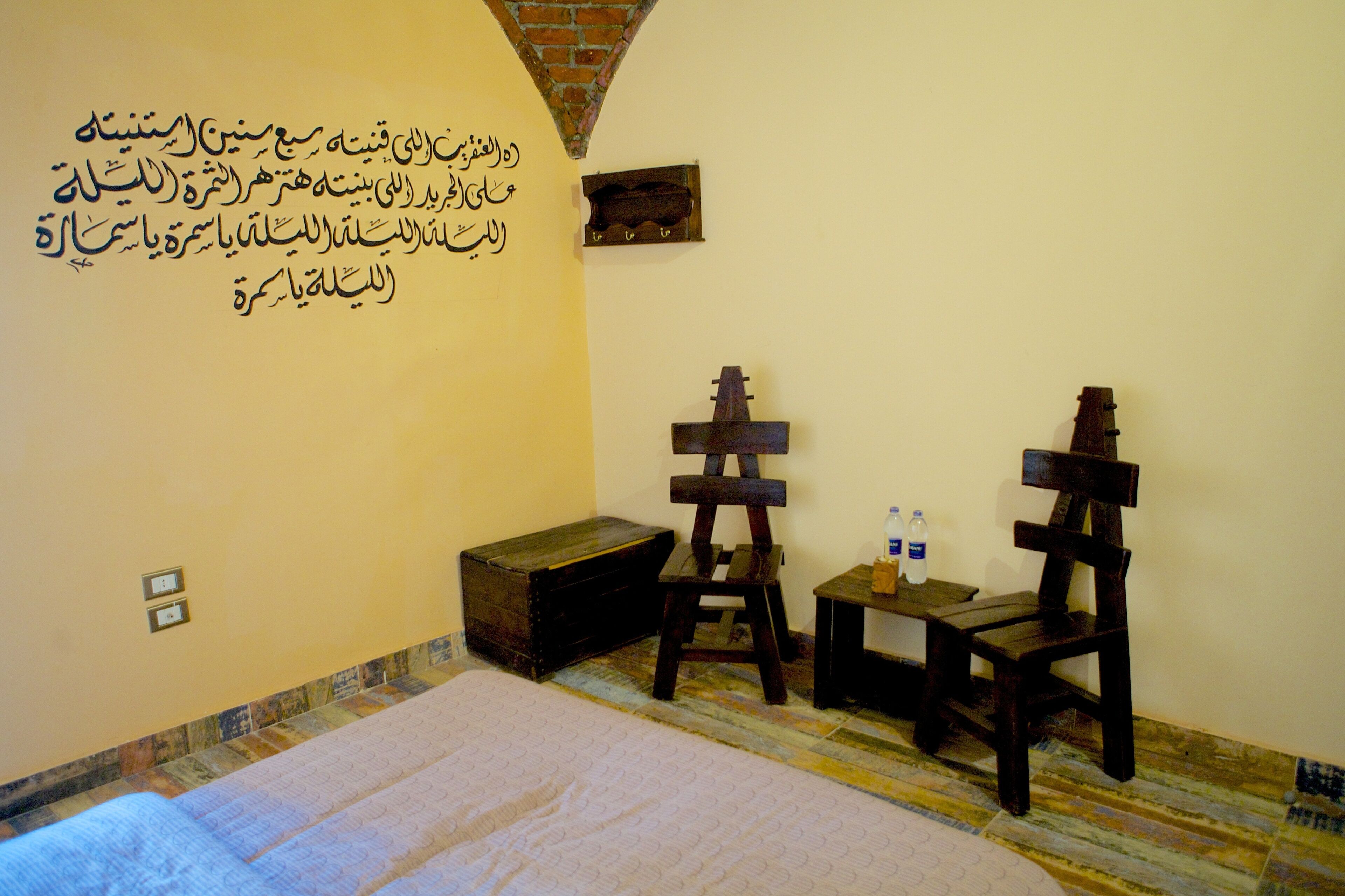 A room with a bed, a wooden chest, two wooden chairs, a small table with bottles, and Arabic calligraphy on the wall.