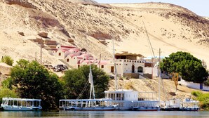 A coastal village with traditional white and red buildings, a large white building with a balcony, and several boats docked at the shore. - Hadouta Masreya - Nubian Guest House (Aswan)
