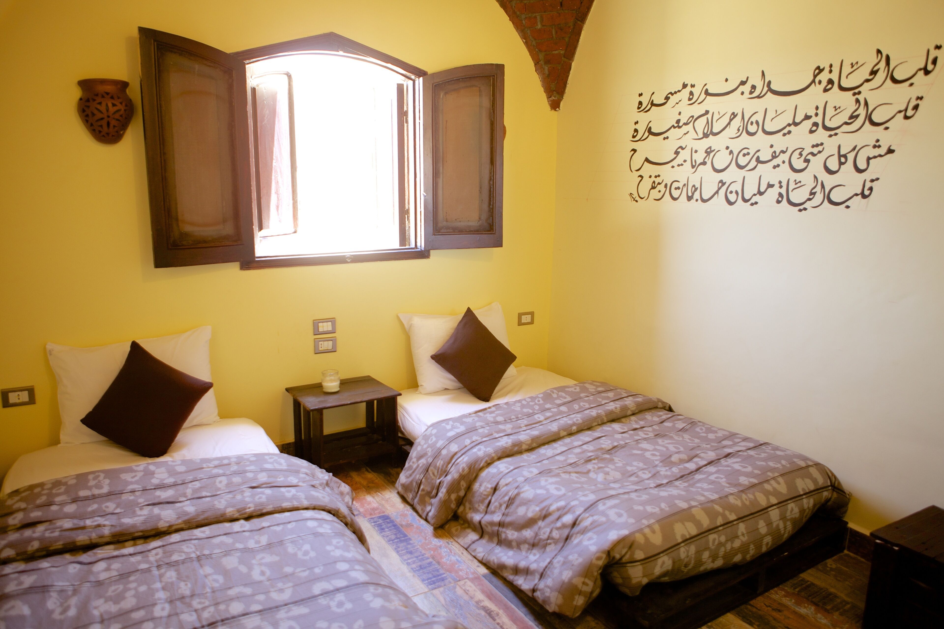 A room with two beds, a window with open shutters, a small table, and a wall with Arabic script.