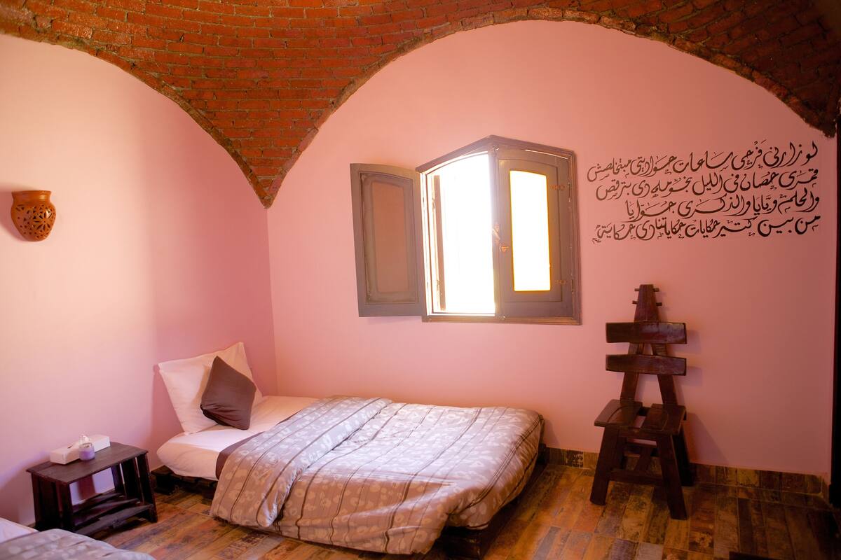 Une chambre avec un plafond en briques, un lit recouvert d’une couvre-lit à motifs, une petite table avec une lampe et une fenêtre donnant sur l’extérieur.