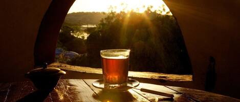 A glass of beer on a wooden table with a view of a sunset through an arched window.