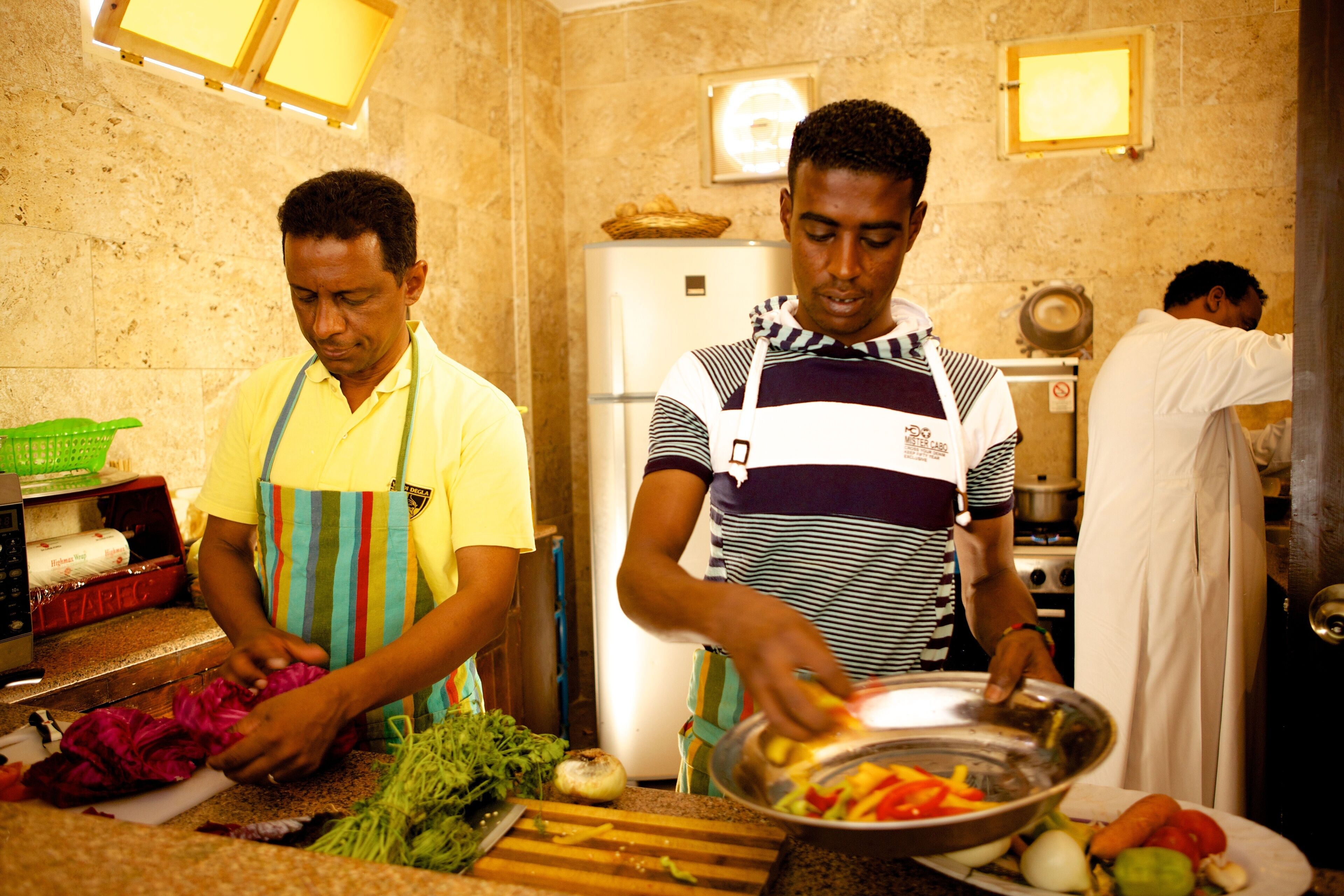 A kitchen with two individuals preparing food, a refrigerator, and various vegetables on the counter.