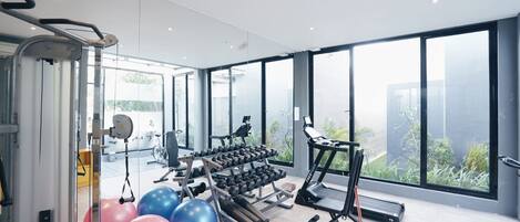 A modern gym with exercise equipment, large windows, and a view of greenery.