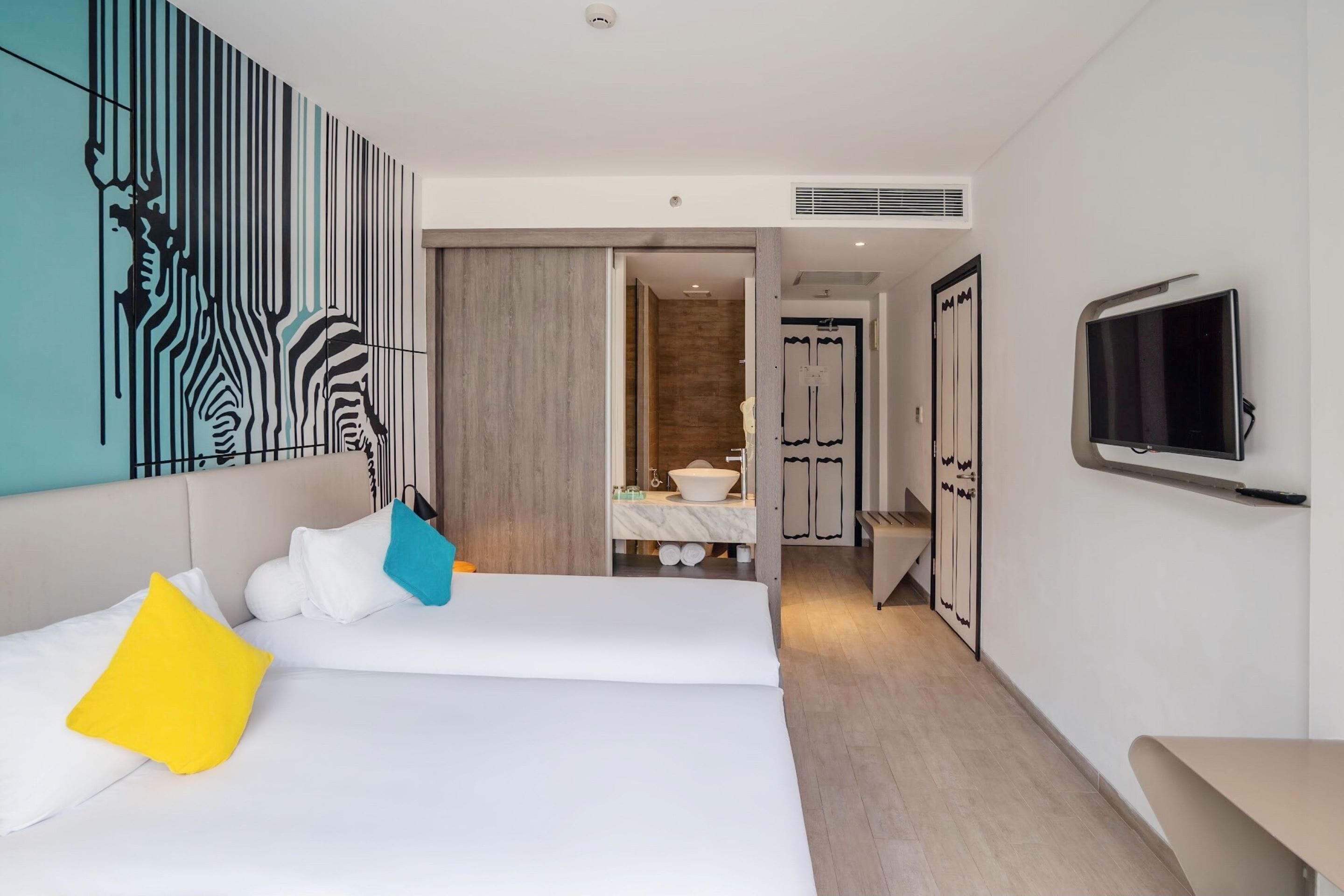 Una habitación de hotel moderna con una cama grande, un televisor, un baño con lavamanos y espejo, y una obra de arte original en la pared.